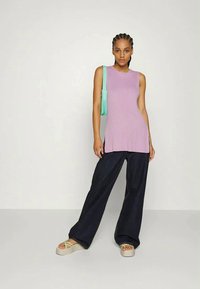 Mulher vestindo uma blusa sem mangas de malha lilás, calças largas escuras, sandálias de plataforma e uma pequena mala de ombro verde-menta.