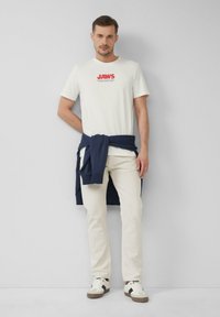 Biała koszulka z grafiką i napisem "JAWS", jasnobeżowe spodnie oraz granatowy sweter przewiązany w talii. Białe sneakersy z ciemnymi akcentami.