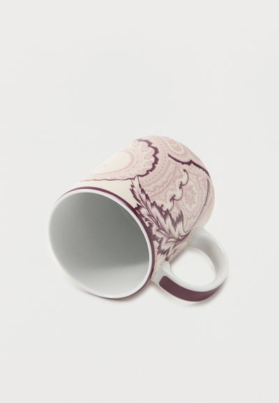 Tasse en céramique avec des motifs floraux et paisley complexes en bordeaux et rose, posée sur le côté avec la anse visible sur fond blanc.