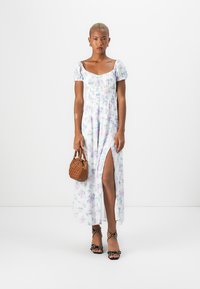 Rochie maxi cu imprimeu floral în nuanțe de albastru deschis și pastel, având mâneci puff, un corset elastic și o fanta pe latura coapsei. Asortată cu o geantă maro.