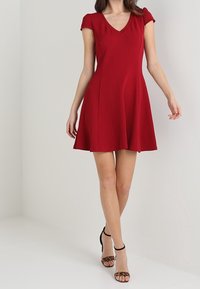 Vestido rojo de manga corta con escote en V, falda acampanada y tejido liso, ligeramente texturizado. Combinado con sandalias de tacón alto negras y con tiras.