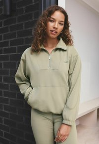 Světle zelený pulovr na zip s vysokým límcem, žebrovanými manžetami a volným střihem. Obsahuje švy a malé logo na hrudi.