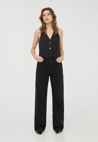 Top nero corto con scollo a V, caratterizzato da tre bottoni e due tasche frontali. Abbinato a jeans neri a vita alta con delicate decorazioni.