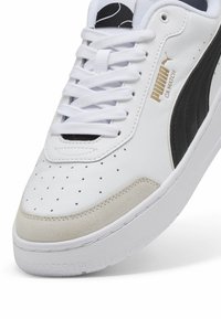 White sneaker featuring a perforated toe, suede detailing, black accents, and gold branding. Lace-up design with a rubber sole.