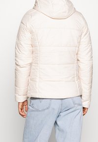 Osoba oblečená v světle béžové prošívané bundě s kapucí a světle modrých džínách, zobrazená zezadu na jednoduchém pozadí.