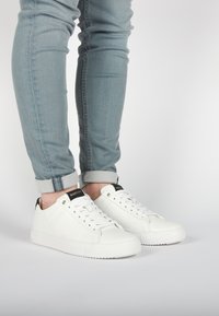 Sneaker bianche in pelle con finitura testurizzata, caratterizzate da dettagli neri sul tallone e sui lacci, abbinate a jeans azzurri chiari con risvolto.