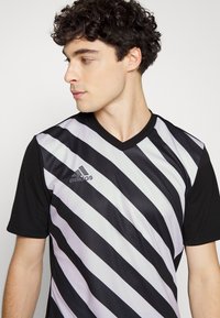 Chemise noire à manches courtes avec un col en V, ornée de rayures diagonales noires et gris clair. Logo Adidas en argent sur la poitrine gauche.