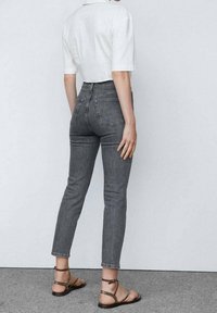 Los jeans grises de cintura alta y corte ajustado presentan una textura de denim clásica y dos bolsillos traseros. Se combinan con una blusa blanca de manga corta y sandalias con tiras.