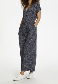 Combinaison florale bleu marine à manches courtes, col en V, taille à nouer et poches latérales, associée à des baskets blanches. Tissu léger.