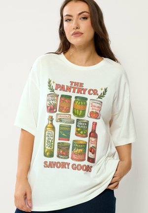 Kvinde iført hvid t-shirt med vintageillustrationer af konservesdåser og flasker samt tekst "The Pantry Co. Savory Goods."