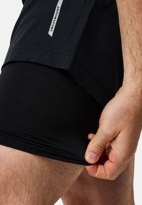 Musta urheilushortsit, joissa on sivulogo, päällekkäin tiukkojen mustien kompressioshortsejen kanssa, joissa on rei'itetty pinta. Näkyvissä käsi, joka säätää kangasta.