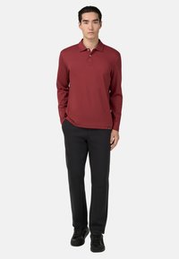 Jaunas vyras, apsirengęs ilgomis rankovėmis bordo polo marškiniais, juodomis kelnėmis ir juodais sportbačiais, stovintis su rankomis iš dalies kišenėse.