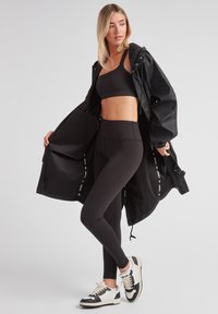Schwarze Sportjacke, offen über einem schwarzen Cropped-Top und hochgeschnittenen Leggings. Die Mode trägt schwarze und weiße Sneaker. Glatte Textur, minimales Design.