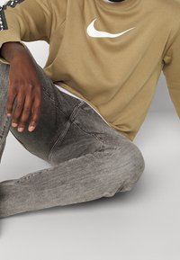 Beige sweatshirt med vitt Nike swoosh-logo, kombinerad med grå denimjeans som har en lätt blekt textur och en klassisk design med fem fickor.