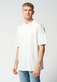 En blond mand iført en simpel hvid oversized t-shirt med korte ærmer og lyseblå jeans, står op af en ensfarvet lys baggrund.