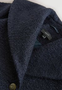 Tumesinist tekstuuriga mantel suure kraega, millel on kuldne nööp ja silt tekstiga "NEXT." Kangas tundub pehme ja paks.