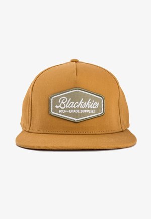 Geelbruine snapback pet met een platte rand en een gestructureerde afwerking. Voorzien van een hexagonaal patch met de tekst "Blackskies" in het wit.