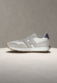 Vue latérale d'une basket gris clair et blanche avec une semelle beige et un contrefort foncé sur une surface beige.