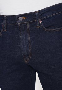 Pantalones vaqueros de mezclilla azul oscuro con costuras naranjas, que presentan un cierre con botón metálico, trabillas para cinturón y un bolsillo delantero. Textura suave, corte clásico.