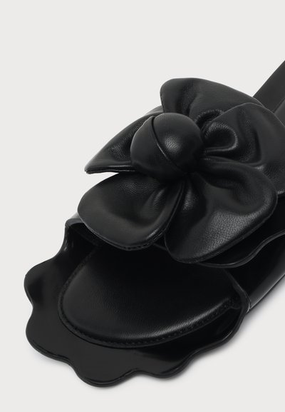 Sandale noire avec une grande fleur en cuir texturé et des bords festonnés sur la semelle, présentée sur un fond blanc uni.