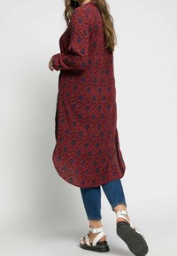 Chemise longue à motif brun rougeâtre avec accents bleus, fentes latérales et manches longues. Associée à un jean en denim et des sandales à brides argentées.