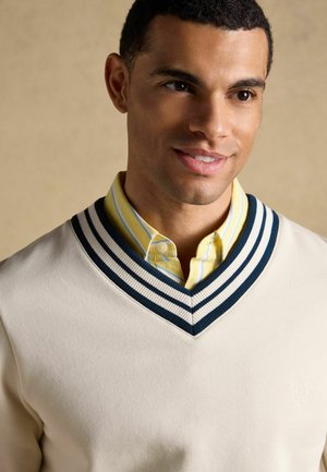 Lächelnder Mann, der einen cremefarbenen Pullover mit marineblauen Akzenten über einem gelb gestreiften Hemd vor einem schlichten beigen Hintergrund trägt.