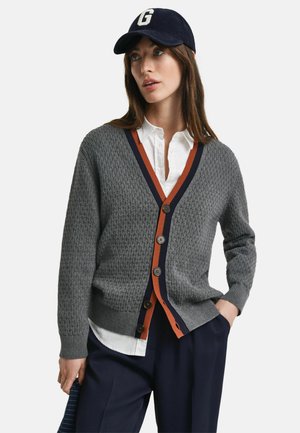 Cardigan gris en tricot avec un col en V, bord orange et marine, fermeture à boutons, porté sur une chemise blanche, associé à un pantalon marine et une casquette sombre.