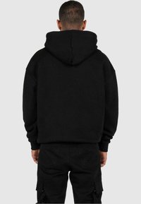 Felpa nera con cappuccio in misto cotone, caratterizzata da una vestibilità larga, polsini a costine e tasca a marsupio, vista da dietro.
