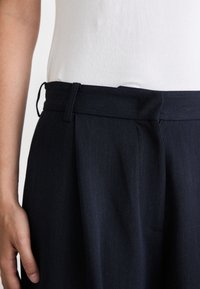 Pantaloni blu navy con una vita strutturata, caratterizzati da pieghe laterali e un passante per cintura in tessuto. Il materiale presenta una sottile trama.