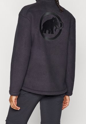 Person trägt eine dunkle Fleecejacke mit einem Mammut-Logo in einem Kreis auf dem oberen Rücken, kombiniert mit einer dunklen Hose.