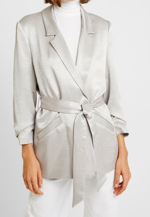 Vrouw draagt een lichtgrijze satijnen blazer met riem over een witte coltrui en witte broek, handen ontspannen langs het lichaam.