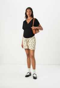 Haut noir enroulé, shorts crème avec motifs noirs, sac à épaule beige, chaussettes blanches et chaussures noires. Le mannequin présente une tenue décontractée.