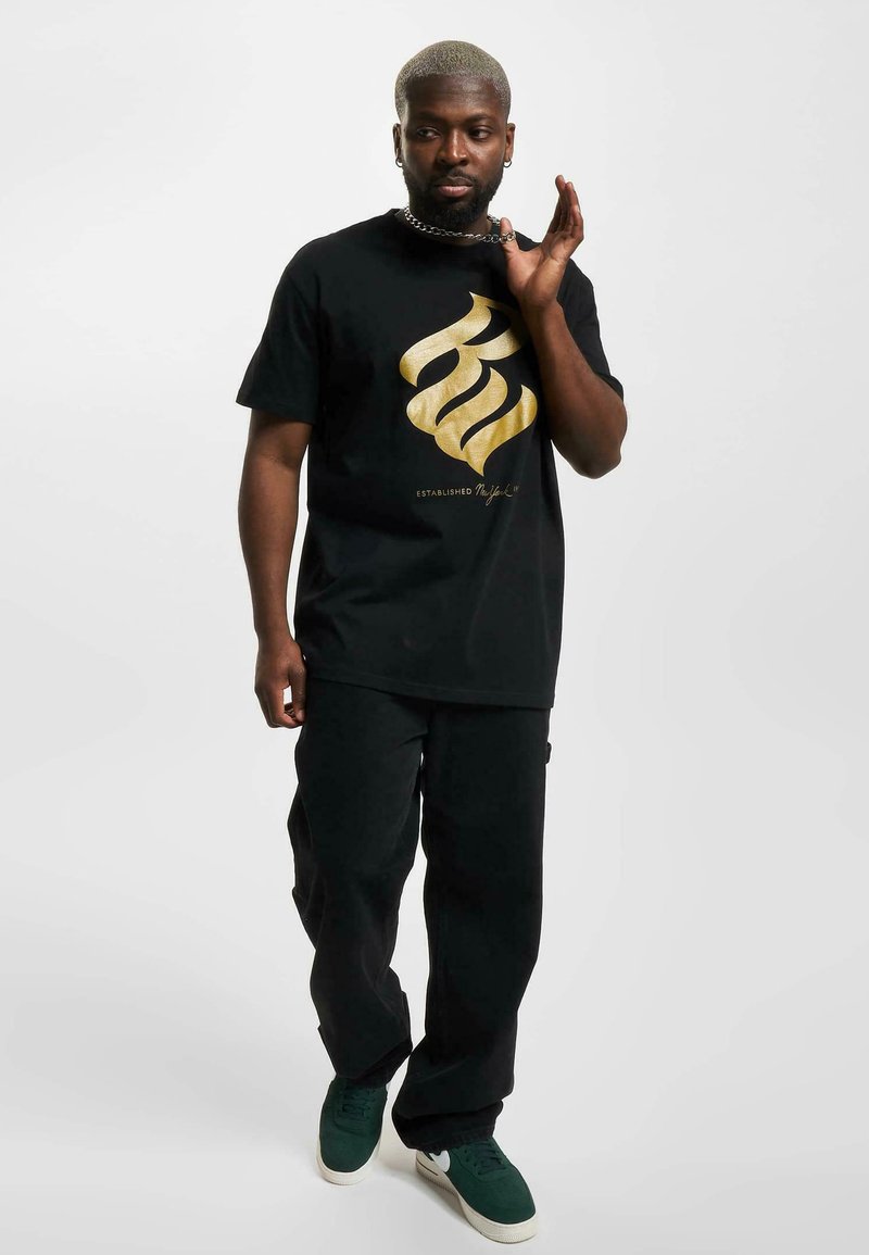Black t-shirt with a large golden graphic design; paired with black pants and green sneakers; short sleeves, relaxed fit.
