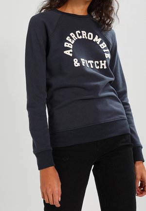 Personne portant un sweat-shirt bleu marine avec le texte "Abercrombie & Fitch" à l'avant et un pantalon noir sur un fond gris.