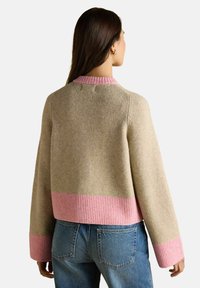 Žena otočená chrbtom, oblečená v béžovom svetri s ružovými rebrovanými manžetami, golierom a spodným okrajom, v modrých džínsoch na pozadí bez vzoru.