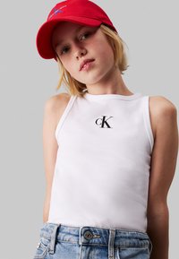 Top bianco senza maniche con logo cK, abbinato a shorts in denim azzurro chiaro. Un cappellino rosso è indossato sulla testa. Design semplice e sportivo.