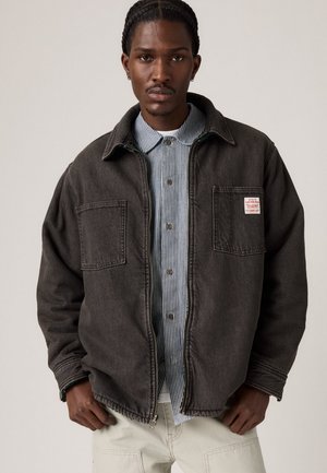Oversized dark gray denim jacket with two front pockets, buttoned collar, and a stitched hem, layered over a striped button-up shirt.