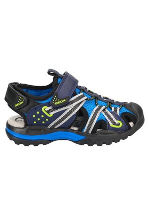 Sandalia azul y negra con parte superior de malla, correa de Velcro y suela de goma. Presenta acentos en verde neón y elementos de diseño a rayas.