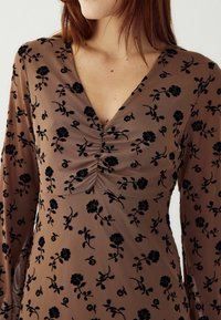 Hnedé šaty so hlbokým V-výstrihom, čierny sametový kvetinový vzor, nariasené detaily na hrudníku a dlhé, voľne padajúce rukávy.