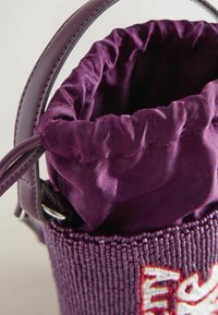 Sac en tissu violet avec cordon de serrage, extérieur orné de perles et poignée en cuir, partiellement ouvert pour révéler la doublure intérieure en tissu froncé.