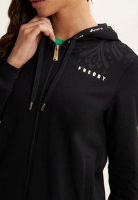Sudadera con cremallera negra y cremalleras doradas, que presenta un diseño impreso en la capucha, el logo "FREDDY" en el pecho y una textura de tela suave.