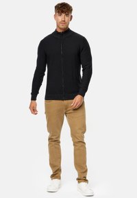 Cárdigan negro con cremallera y textura acanalada, con un cuello alto. Combinado con pantalones beige ajustados y zapatillas blancas.