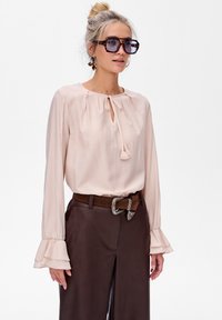Jasnoróżowa bluzka z okrągłym dekoltem i detalem w postaci kokardy, z długimi rękawami zakończonymi falbankami. Połączona z brązowymi spodniami z wysokim stanem i paskiem.