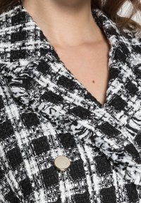 Svart- och vitrutigt blazer tyg med en texturerad yta, som har en guldkalafon på kragen. Tjock väv med synliga sömdetaljer.