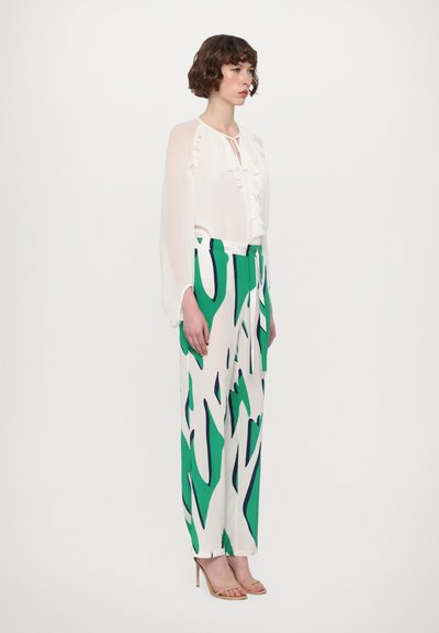 Blouse blanche avec col à volants, associée à un pantalon à jambes larges à motif vert et blanc. Le mannequin porte des talons couleur beige. Texture du tissu lisse.