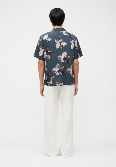 Homme debout de dos, portant une chemise à manches courtes bleu foncé avec de grands imprimés floraux blancs et noirs, et un pantalon blanc à jambes larges.