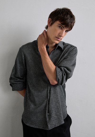 Camisa de botón gris con patrón, cuello, mangas enrolladas y una tela sutilmente texturizada. El modelo posa con una mano en el cuello contra un fondo neutro.