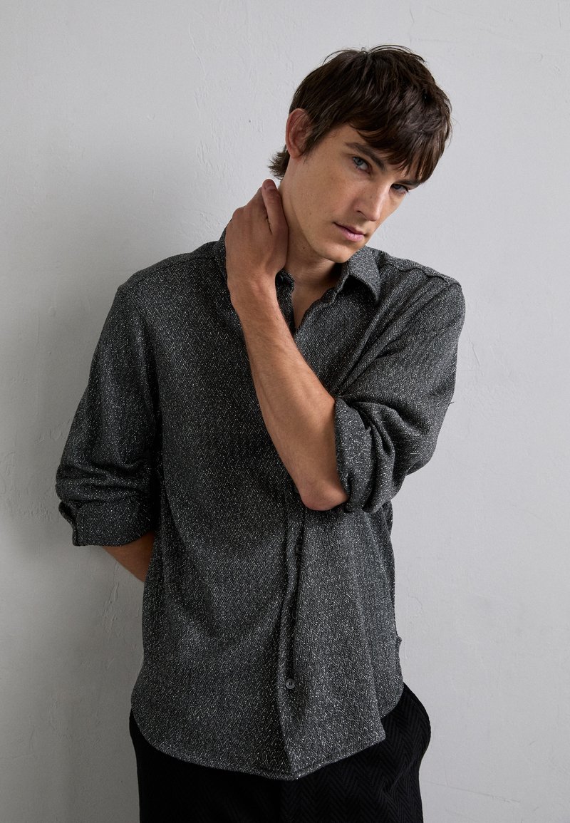 Gråmønstret skjorte med knapper og krave, opkrøllede ærmer og et subtilt struktureret stof. Modellen poserer med den ene hånd på nakken foran en neutral baggrund.