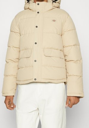 Beżowa pikowana kurtka puchowa z zatrzaskami i klapami na kieszeniach, noszona z białymi spodniami, ręce swobodnie ułożone wzdłuż ciała.