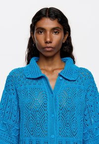 Cardigan lavorato a maglia di un blu vivace, con colletto, chiusura a bottoni e intricati motivi a giorno in tutto il tessuto.